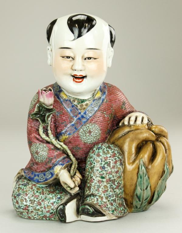 CHINESE PORCELAIN FIGURE OF A CHILD: In seated position and wears a colorful multi-pattern robe. He leans against a large citron fruit and holds a long stem lotus flower. H: 11 1/8 in.