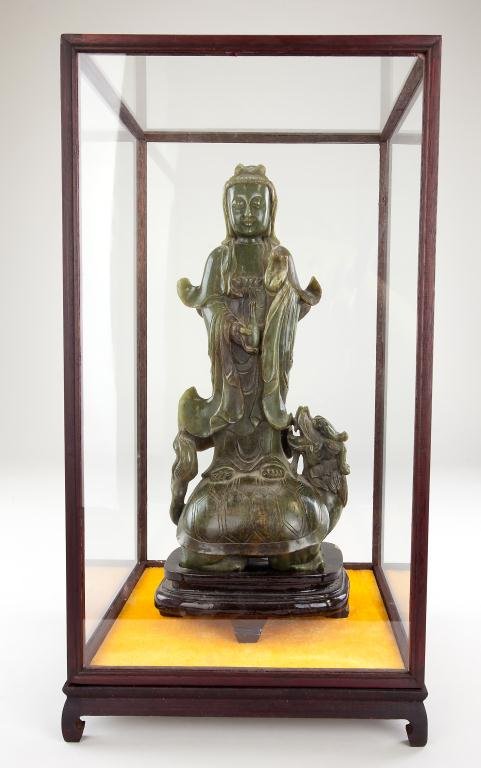 CHINESE CARVED SPINACH GREEN SOAPSTONE GUANYIN: CHINESE CARVED SPINACH GREEN SOAPSTONE GUANYIN Standing atop a turtle dragon, her long robes floating up on the sides. She holds a water vessel in her right hand at her front. On wood stand, in glass 