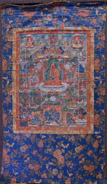 TIBETAN THANGKA ON TEXTILE: Centrally painted with a Buddha on a lotus throne, surrounded by smaller deities. Mounted on embroidered textile with blossoming peonies against a blue background. 24 1/2 in., L: 17 1/2 in.
