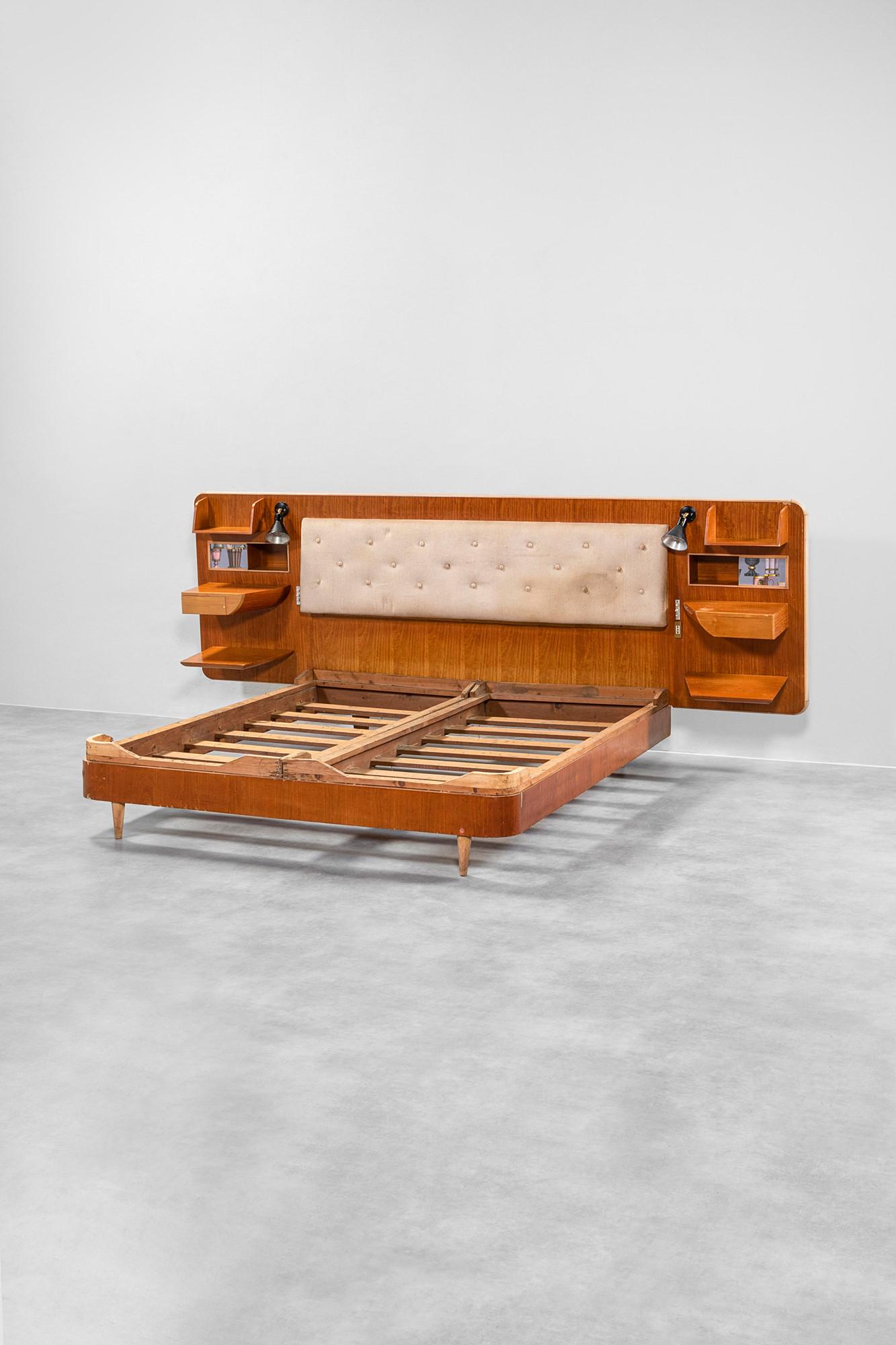 Gio Ponti: Letto con testiera attrezzataTestiera composta da: quattro mensole, due comodini, due vani portaoggetti con portafotografie integrato.Legno di acero e di pero, tessuto Fornasetti, illuminazione non co