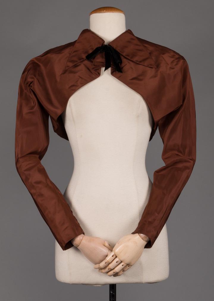 CHARLES JAMES EVENING BOLERO, 1952: Bolero jacket in cocoa brown silk taffeta w/ decorative black velvet ribbon at jewel neck, extended lobed tab collar above fluted jabot to slanted cut-away & long tapered sleeves w/ 5" zippers at insi
