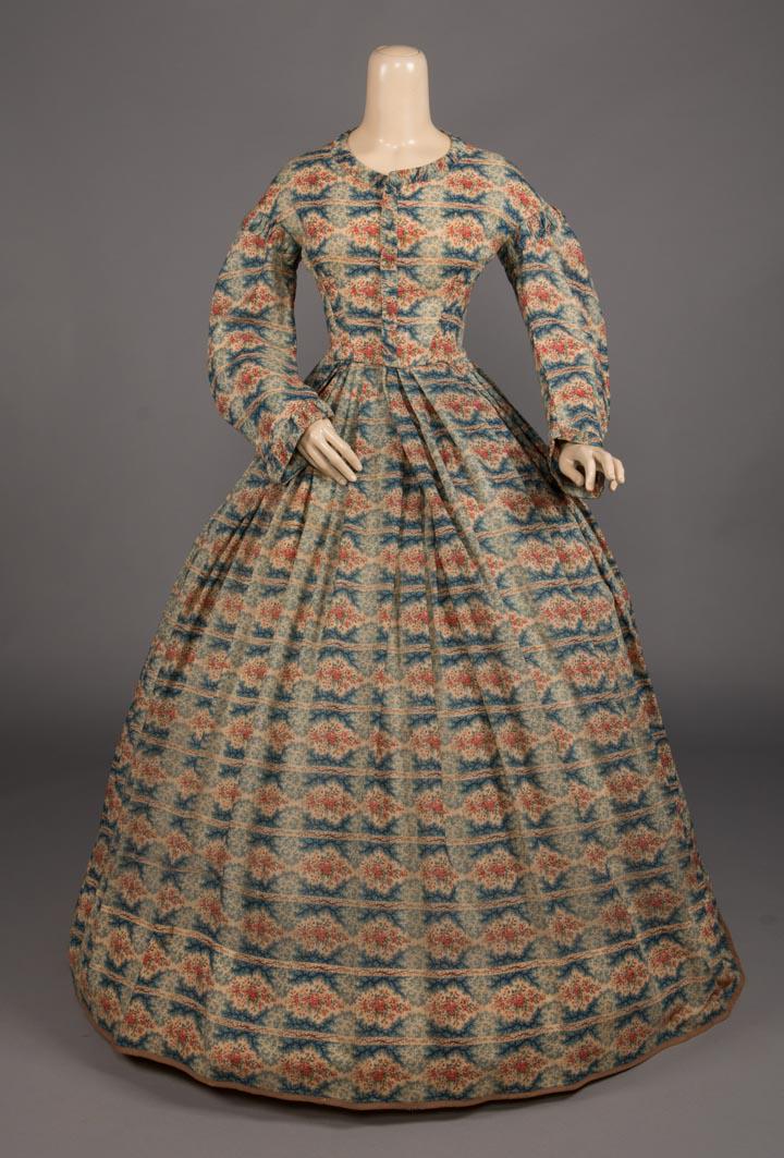 PRINTED WOOL GAUZE DAY DRESS, c. 1863: Day dress in printed blue, red & white floral spray interspersed w/ blue & white abstract branch motif & brown stripes w/ CF closure, gathered detail at collar, low SH & cuffs, wide box pleated skirt,