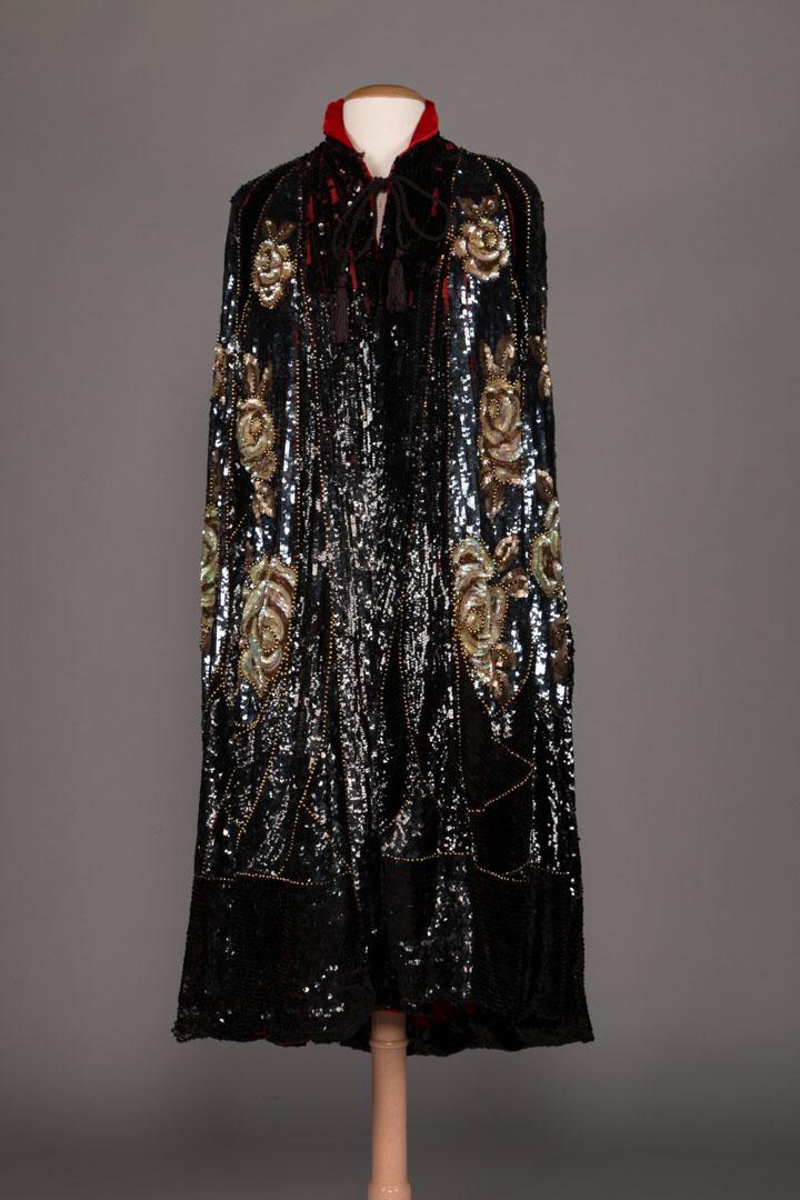 SEQUIN ENCRUSTED OPERA CAPE, 1920s: All over black, navy & oppalescent sequins in rose motif rimmed in gold ball beading, lined in crimson orange velvet, CF cord closure, CB L 49", (sequin & bead loss, tear in sequined fabric at back of