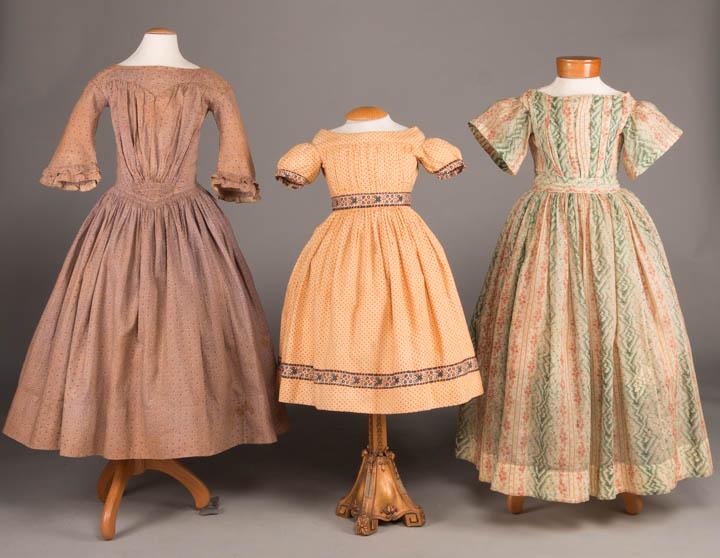 THREE GIRL'S DAY DRESSES, AMERICA, 1840-1850s: 1 brown resist dyed roller printed cotton w/ gathered shirred B to upper yoke, bell sleeves, piping throughout, 1 H pocket, c. 1845, CH 23", W 21", L 36.5", (stains on SHs, wear spots to neckline, sta