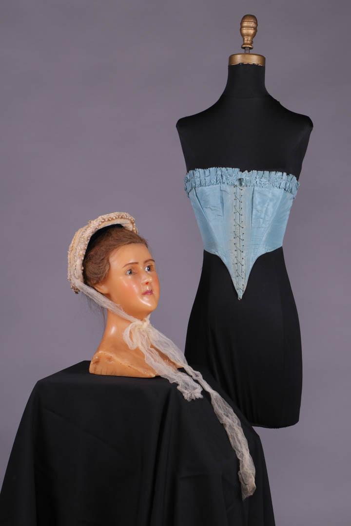 SILK CORSELET & WEDDING BONNET, c. 1855: 1 blue silk taffeta corselet, CF lace closure w/ thread eyelet over boned edge, deep V point at CF & CB, upper edge finished w/ double box pleated ruffle w/ pinked edge, part of wedding trousseau, sea