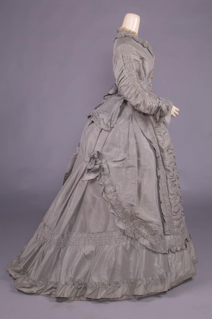 GREY SILK TAFFETA PROMENADE GOWN, c. 1869: 2-pc elephant grey silk taffeta gown, fitted bodice w/ deep pointed front & tabbed back peplum, CF thread covered needlelace button closure, four rows of reverse ruching to outside of full gathered sl