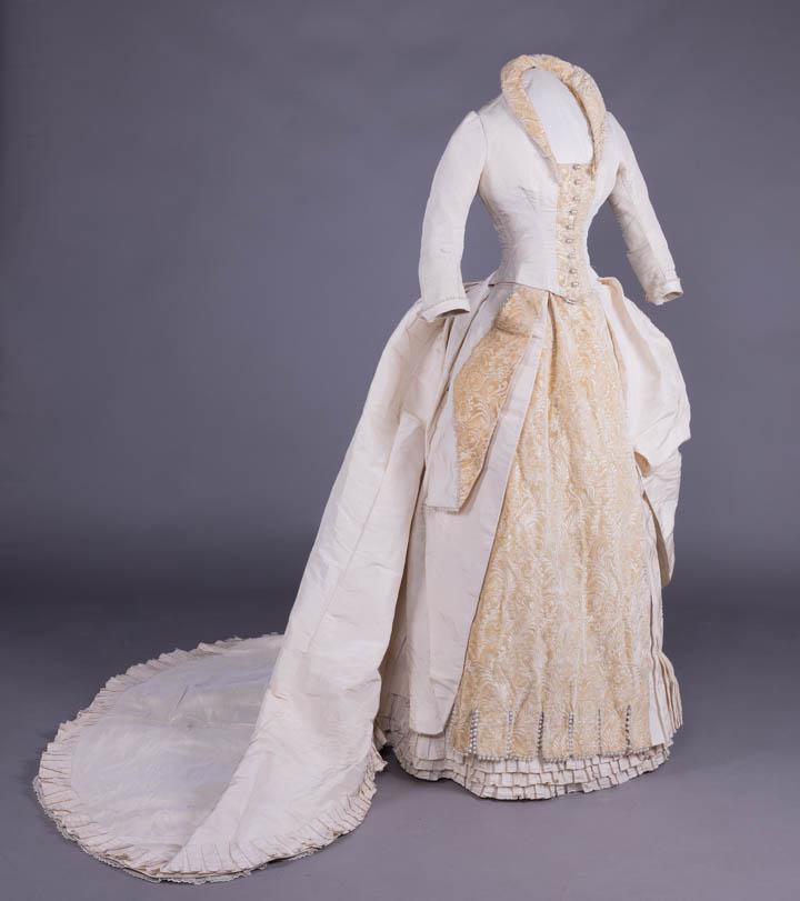 BROCADED VELVET & SILK FAILLE WEDDING GOWN, c. 1883: Bone silk faille wedding gown w/ brocaded velvet collar, CF placket & skirt panels, trimmed w/ faux pearls, asymmetrically draped & swagged princess line train w/ box pleated hem & mm lace hem flounce