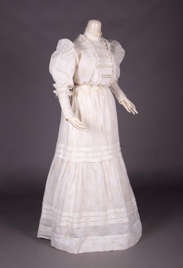 COTTON DAY DRESS, c. 1910: 2-pc white cotton day dress printed w/ overall sprig motif, horizontal tucking & lace insertions to plastron front & deep extending cuffs, 2- tiered skirt w/ double row of triple tucks B 42.5", W 40.5