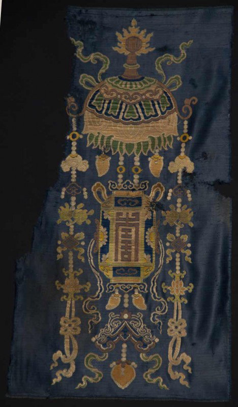 SILK BUDDHIST UMBRELLA FRAGMENT, TIBET, 17TH C: Brocade textile woven w/ umbrella above other religious iconography in shades of ivory, green, gold, blue & brown, dark blue satin ground, irregular 16" x 30", woven in China for Tibetan market, (hole