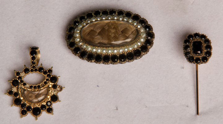 TWO PIECES HAIR MOURNING JEWELRY, c. 1830: Both gold w/ plaited light brown hair under glass surrounded by faceted jet stones: 1 oval brooch, seed pearl row next to jet, inscribed "E.K.H. from Jane", 1.20" x 1.75", excellent; 1 small starburst