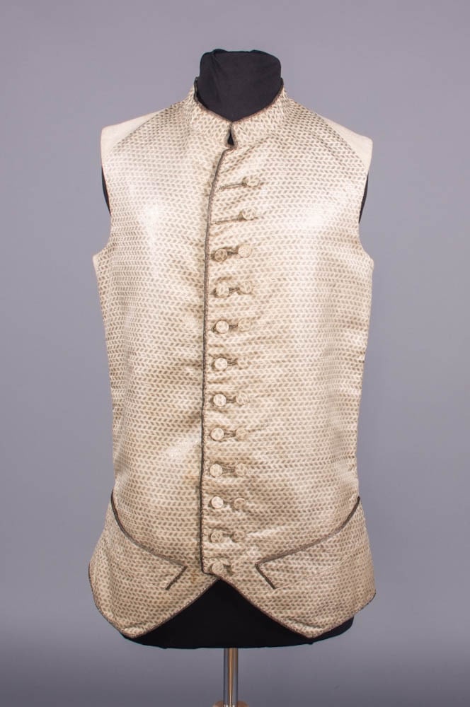 GENTS METALLIC & SILK WAISTCOAT, c. 1765-1780: Man’s waistcoat woven w/ metallic weft interrupted chevron motif, short standing collar, paired CF button closure, warp face silver ribbon edging, fustian lining & backing, CH 36”, L 28”