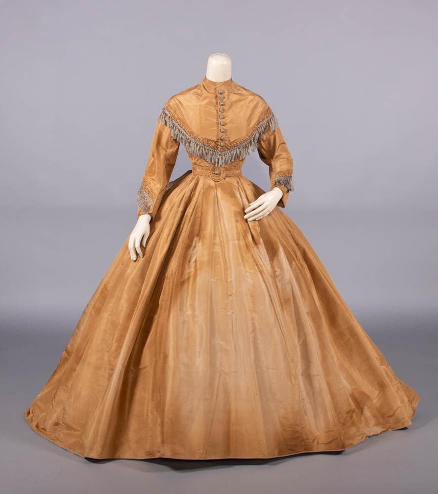 SILK TAFFETA DAY DRESS, 1867: Day dress of Bismarck brown silk trimmed w/ blue-grey silk fringe & covered buttons, trained attached skirt lined w/ glazed cotton, matching self belt w/ panache to CF & flat bow to CB, 1867, good-ver