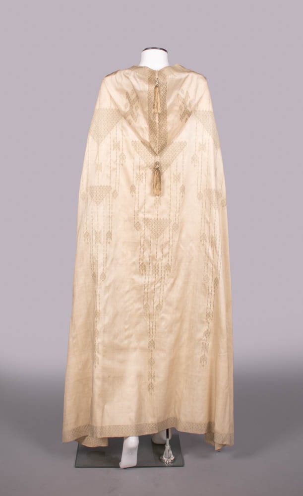 BROCADED SILK & LAME’ BURNOOSE, c. 1867: Cream silk & lame’ burnoose woven to shape in one piece w/ brocade silver lame’ threads, attached hood w/ 2 lame’ & silk tassels, L 56”, (slip N/I, discoloration w/ light staining up to 7.