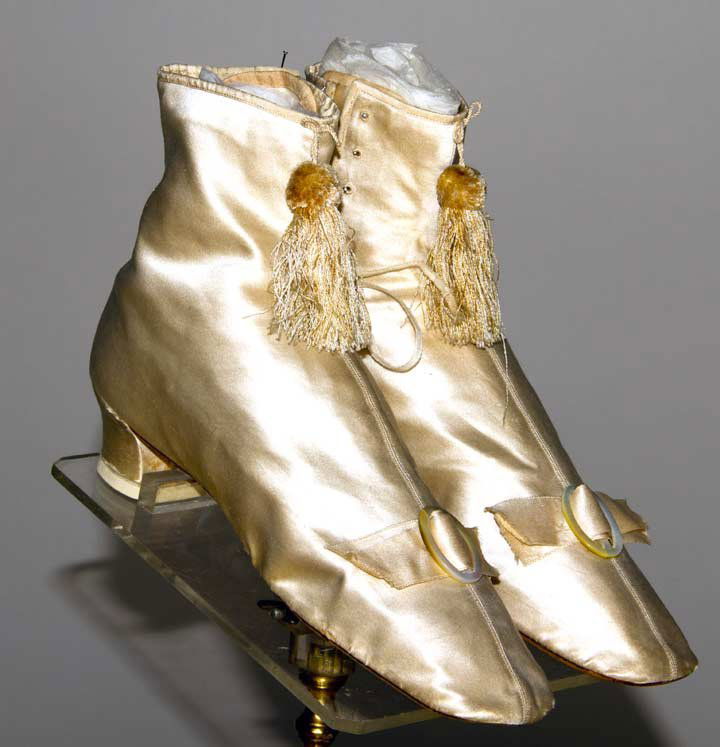 PAIR WEDDING BOOTS, 1840-1850s: White silk satin straights w/ short heels: 1 pair side lace boots, buckle on vamp, tassel on tops, leather marked on soles "3 1/2 571 M J. Eppert New York", (originally slipper boots, heels added late