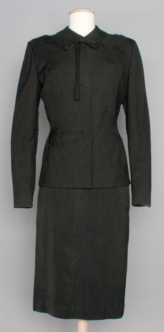 ADRIAN BLACK SKIRT SUIT, 1940s: Wool gabardine, jacket pieced w/ curved panels, 2 pocket flaps just below each shoulder, self covered buttons, string bow at collar, labeled, B 38", W 27", Jacket CB L 25.5", Skirt L 28", excellent.