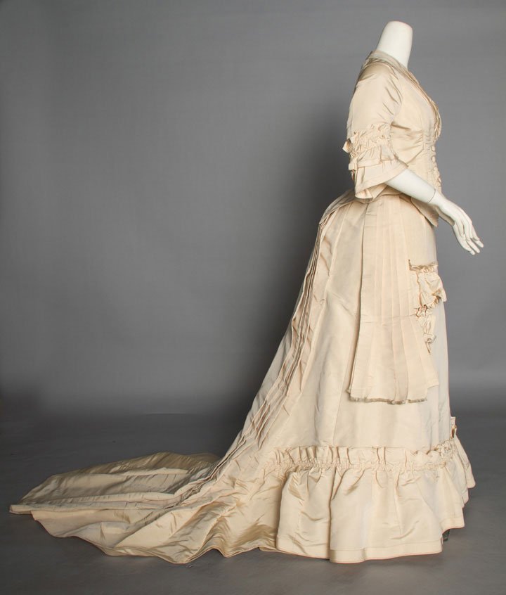 CREAM SILK WEDDING DRESS, 1875: 2-piece faille, elbow length sleeves, trained skirt w/ deep hem ruffle, knife-pleated sashes, self fabric "wedding pocket", skirt lined w/ buckram, B 34", W 22", Skirt L 39"- 69", (hem edges soiled) e