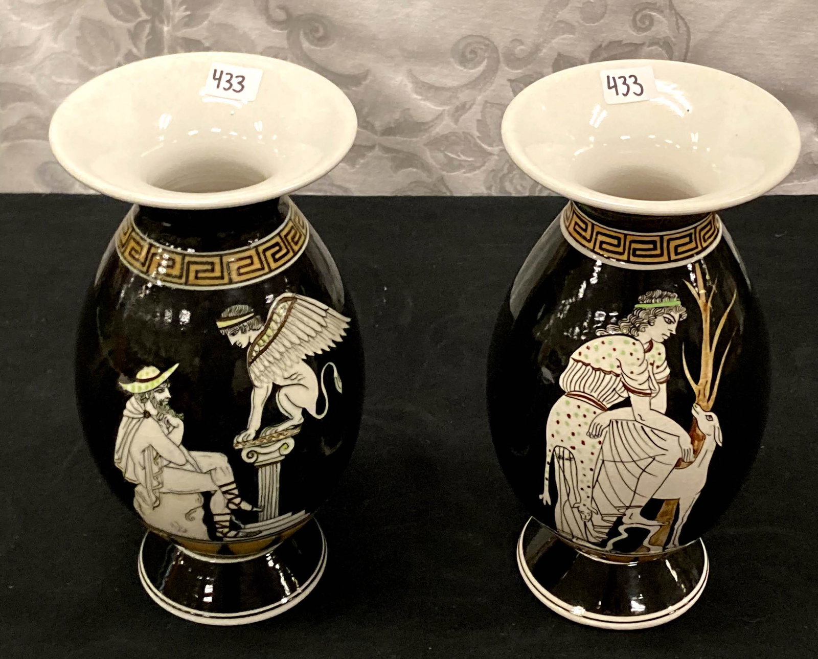 Pair of Greek Art Urns: A pair of Greek art porcelain urns, one depicting a man and a sphinx, and the other depicting a lady and a deer. They measure 9" tall X 4.5" across.