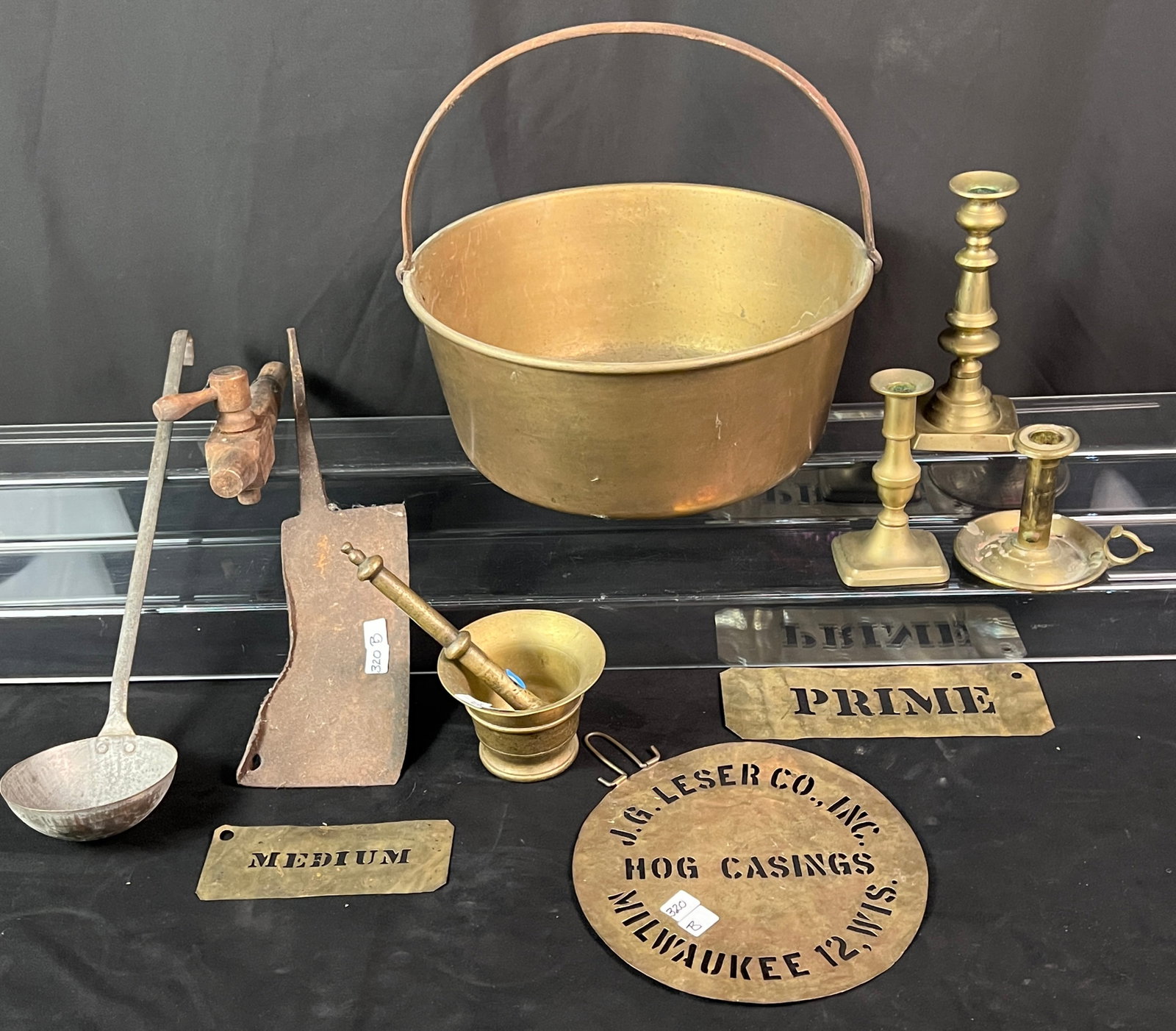 A Large Apple Butter Bucket, Brass Candle Sticks, and More: A group of antique cooking utensils to include a brass bailed handled apple butter bucket. Three early brass push up candle sticks. A mortar and pestle. a brass stencil for J.G. Leser Hog Casings Milw
