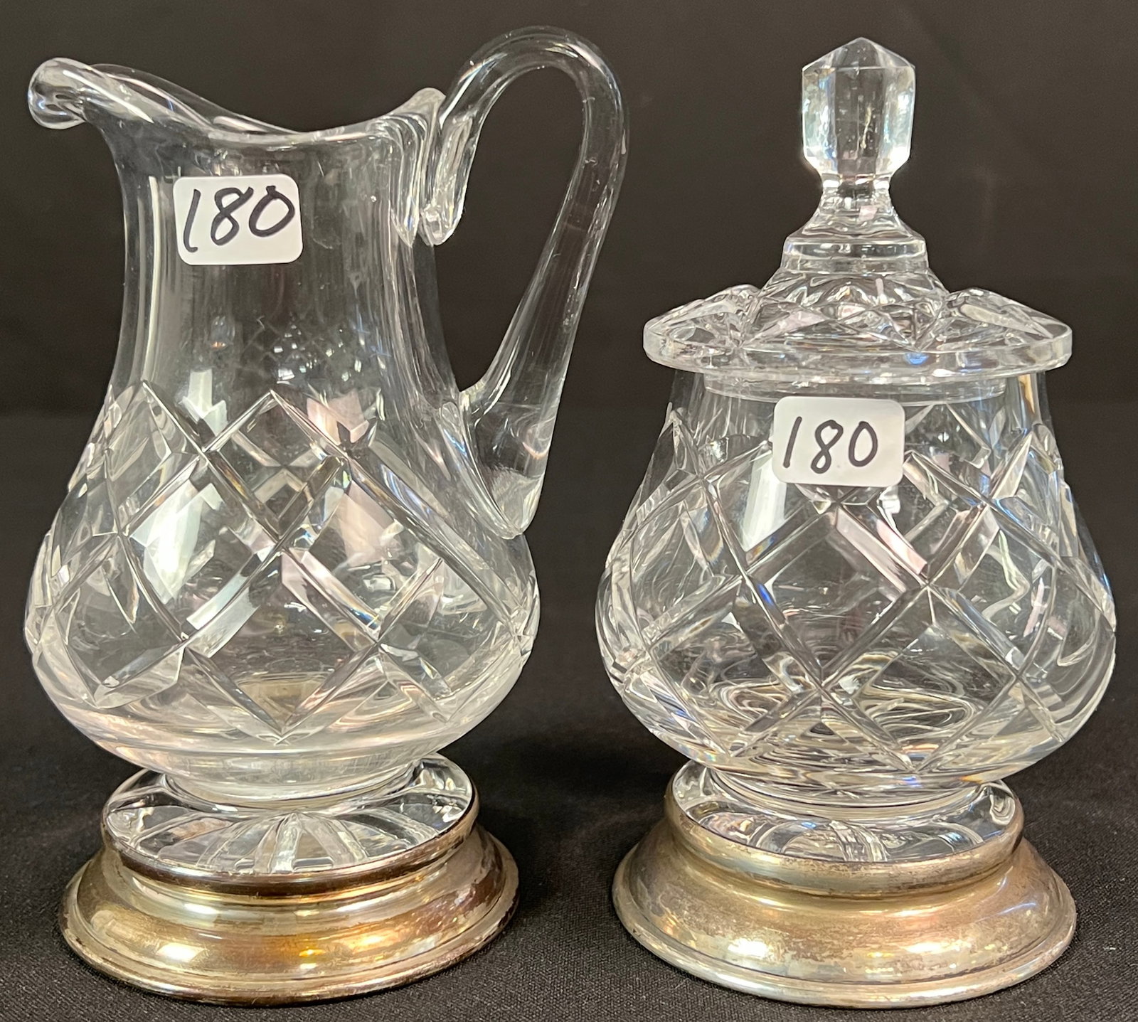 Waterford and Sterling Base Creamer and Sugar: A vintage Waterford creamer and lidded sugar jar, likely from the Alana collection, featuring diamond-cut patterns. Both pieces are set on a sterling silver base. The creamer measures 5.5" tall X 3"