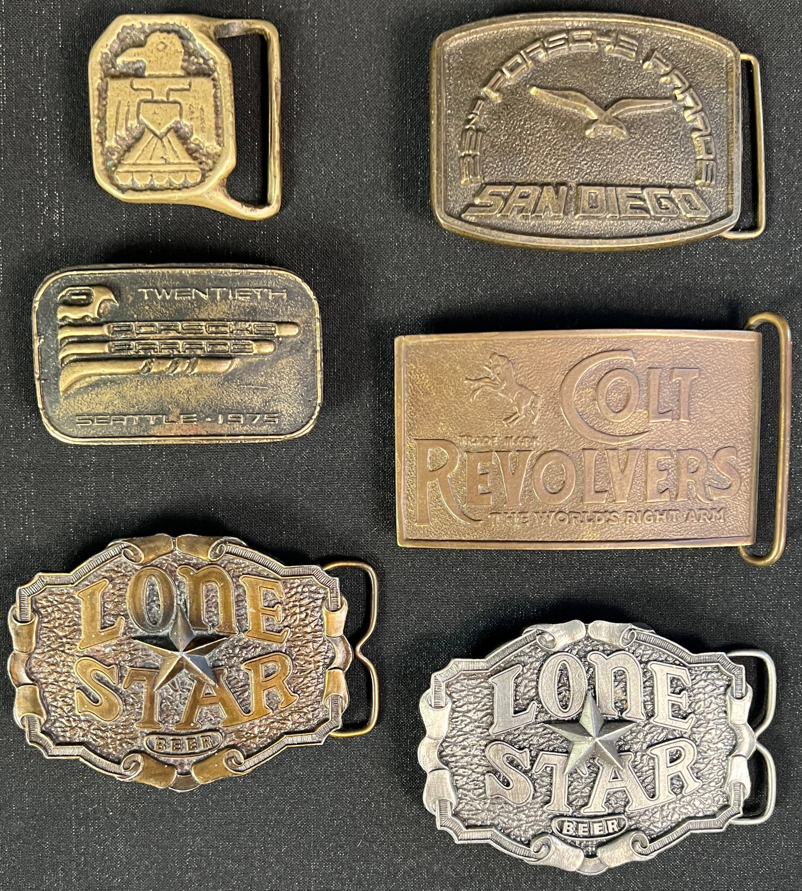 6 Large Belt Buckles: A group of 6 large belt buckles to include a bronze Lone Star Beer buckle, a bronze Porsche Parade Seattle buckle, a bronze Colt Revolver buckle, a bronze Porsche Parade San Diego buckle, a brass hand