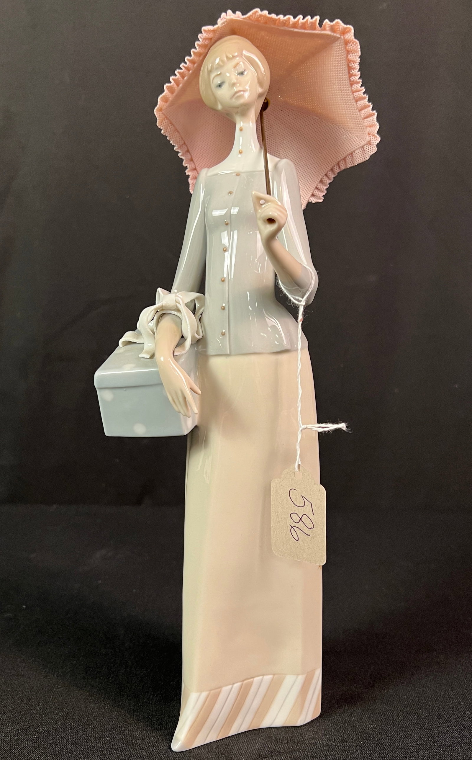 Lladro Porcelain Figurine: An Lladro porcelain figurine titled "The Dressmaker" featuring a lady with an umbrella and gift box. In great condition with minor loss to the umbrella, it measures 14" tall.