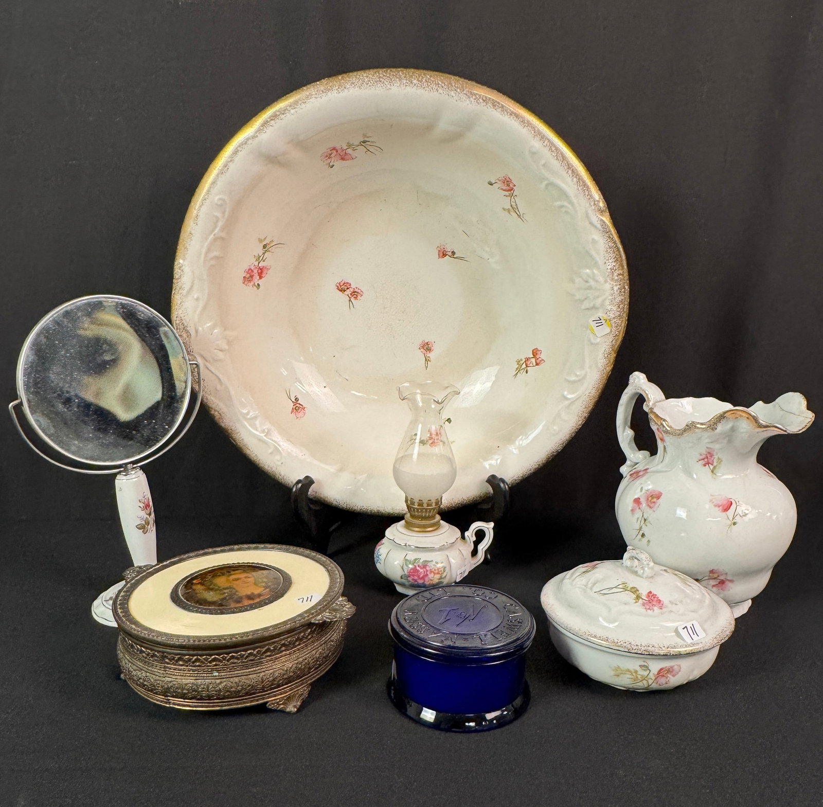 Group of China Dresser Items and Grooming Accessories: A vintage group of China washing and grooming items to include a large floral wash basin, small 8" tall water pitcher, a covered soap dish, a miniature oil lamp, a standing mirror, a covered metal