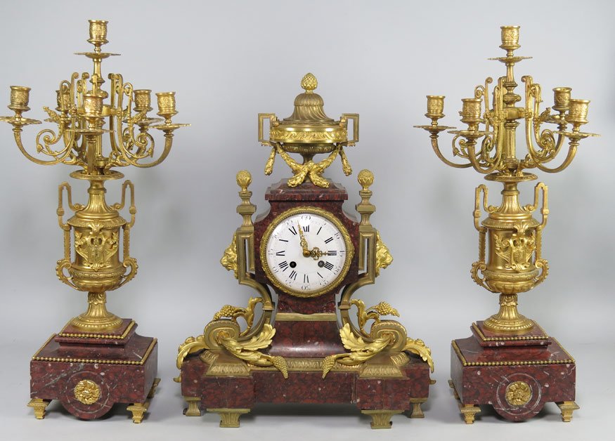 FRENCH ROUGE MARBLE AND FIREGILT BRONZE CLOCK SET: 19th c., sgn. illegibly on porcelain dial ?...RMS..?, the clock, 22”h; 16”w; 7”d; the pair urn-form firegilt garnitures with five scrolling arms centering a candle holder, mounted on marble base