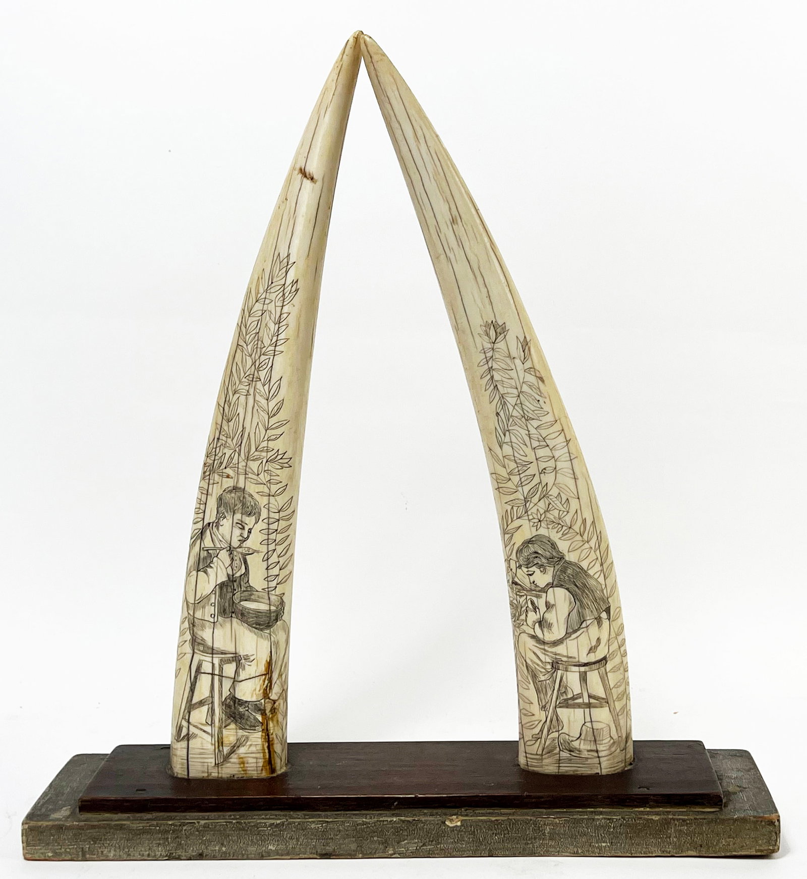 PAIR OF 19TH C. SCRIMSHAW WALRUS TUSKS: one decorated with boy eating porridge sitting amongst foliage; the other, a boy seated in foliage feeding birds, reverse undecorated; tusks measure 15.5"h; mounted on old wood base 16 by 4 in. NOTE: 