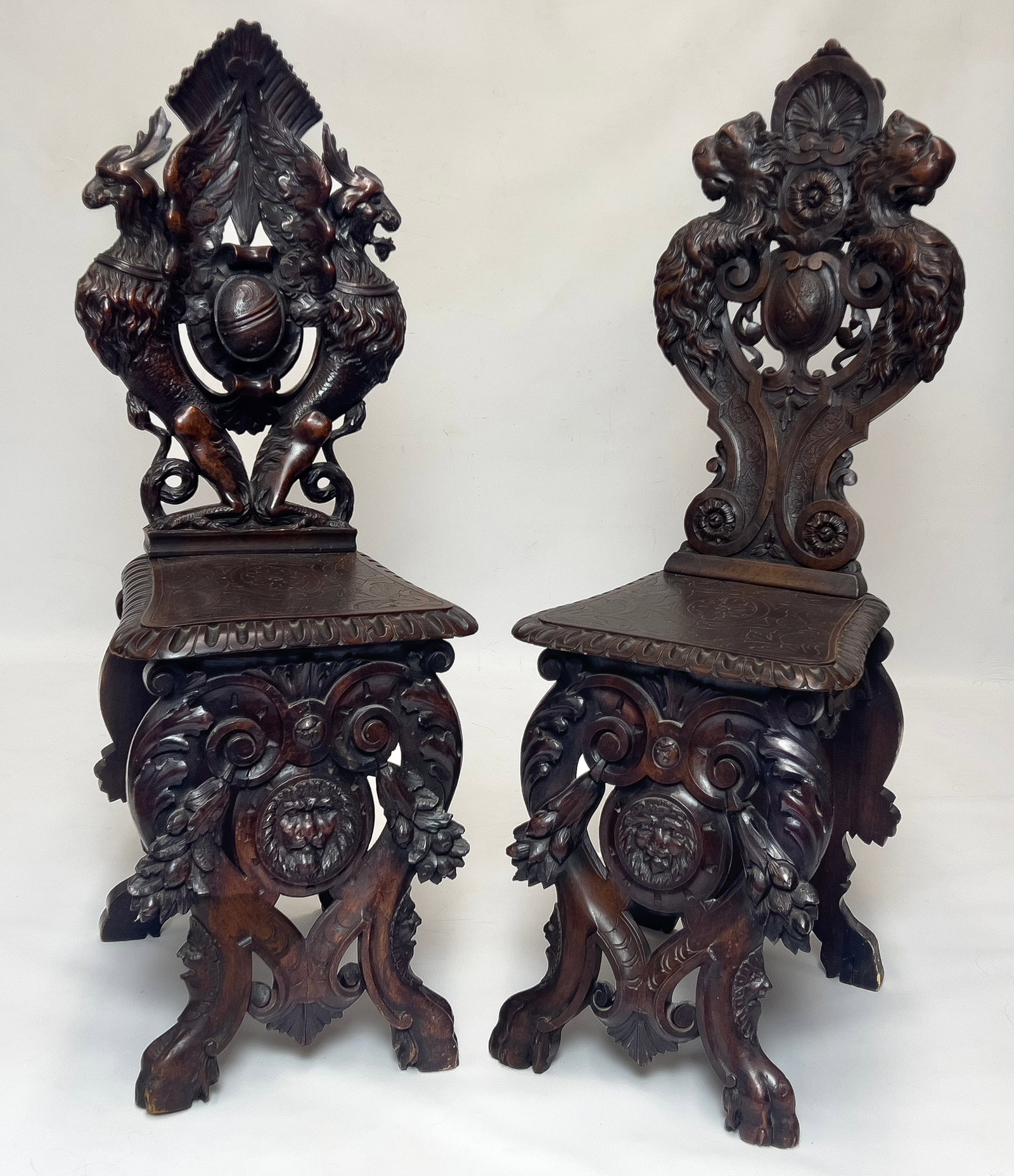 NEAR PAIR OF ITALIAN DEEPLY CARVED SGABELLO CHAIRS: 19th c., Renaissance-style; ornately carved backs, one with pair of lions, the other with winged dragons centering family crests; bold paw feet; 45"h; 15.5"w; 18"d; 20.5" seat height. Carved by the sa
