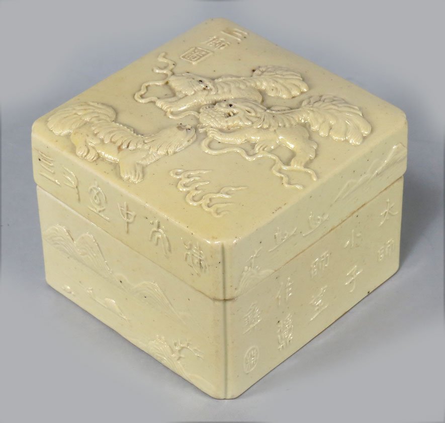 CHINESE PORCELAIN COVERED BOX: square-form, lid with raised decoration of three foo dogs in clouds, raised calligraphy on two sides alternating with lake landscape, bottom with six-character raised mark, 2.5”h; 3.5”square