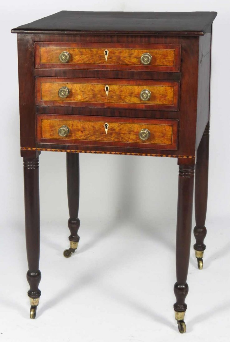 SHERATON FLAME BIRCH AND MAHOGANY STAND: North shore, 3-drawers with flame birch fronts, brass knobs and ivory escutcheons, turned legs with brass cup casters, 31”h; top 18”square