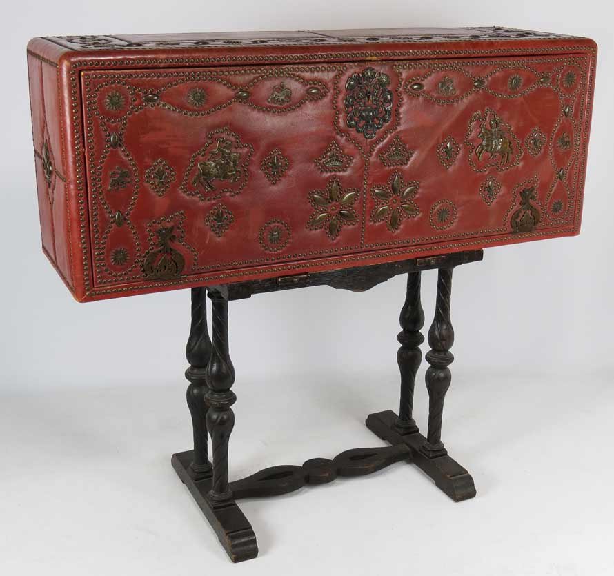 MOORISH-STYLE TOOLED LEATHER FALL FRONT DESK: overall brass studs, red leather, lid with applied brass soldier on horse, interior fitted with drawers and compartments, 19.5”h; 50”w; 15.5”d; on an antique carved wooden stand (not original), 