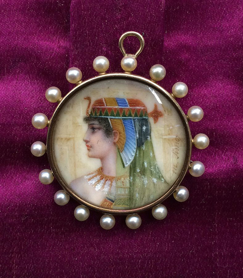 EGYPTIAN REVIVAL GOLD PENDANT/PIN: c. 1872, sgn. M. Sims, miniature profile portrait on ivory of Egyptian princess, in gold frame set with seed pearls, 1.25”diam. Artist Mary Sims was the niece of, and trained with, portrait painter