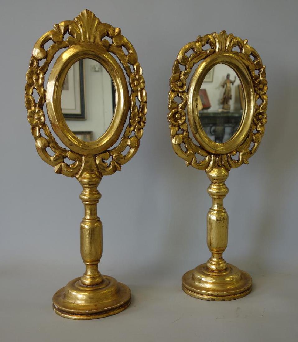 2 Chinese Carved Gilt Wood Table Mirrors: Two similar Chinese carved wood and gilded table mirrors. The oval mirrors framed by rosettes and leaves. Measurements: 20.5" and 22" high, 5.75" diameter at the base.