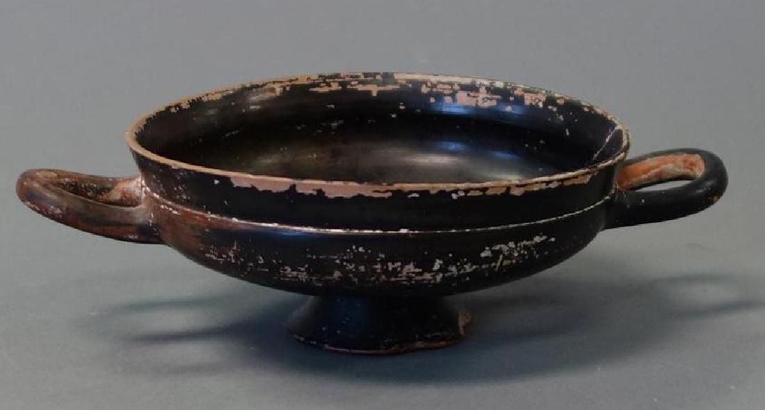 Ancient Greek Pottery Skyphos Wine Cup: An ancient / antique Greek pottery skyphos (two-handled wine cup). Black / brown glaze skyphos on a low flanged footed base with two loop side handles. Measures 7.25" wide to end of handles, 4-5/8" di