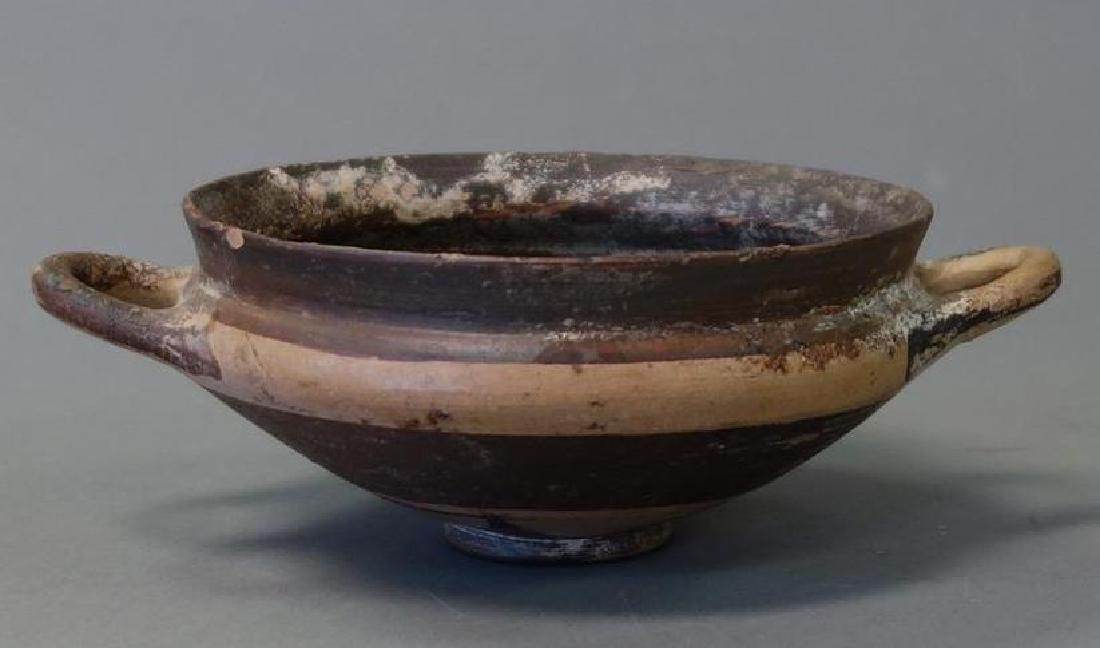 Ancient Greek / Etruscan Pottery Skyphos Wine Cup: An ancient / antique Greek / Etruscan pottery skyphos (two-handled wine cup). Tan brown glaze skyphos on a low flanged footed base with two loop side handles. Measures 6-3/8" wide to end of handles, 4