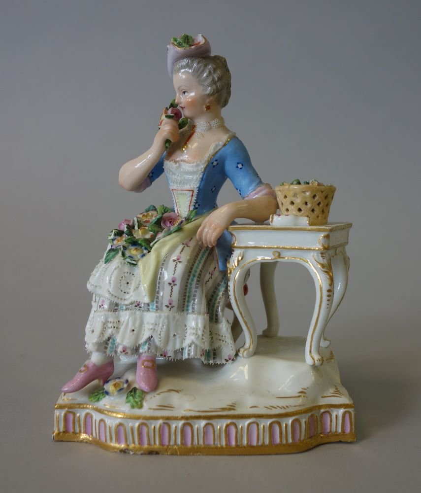 Meissen Porcelain Figurine, The Senses, Smell: A fine Meissen porcelain figurine from the Senses set. Titled "Smell" which depicts a young woman smelling a bouquet of flowers. A basket of flowers and a small vase rest on the side table. Modeled by