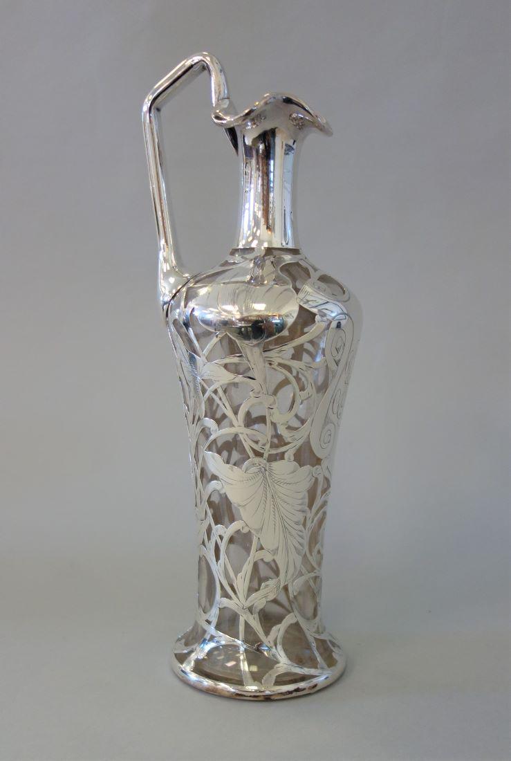Art Nouveau Sterling Overlay Ewer / Wine Pitcher: An Art Nouveau sterling silver and glass ewer / wine pitcher. Graceful sterling overlay of flowers / calla lilies, leaves and scrolling vines. Indistinct maker's mark at lip. Stylized three-initial mo