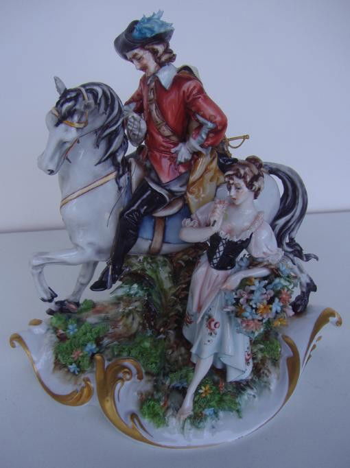 Capodimonte Porcelain Figural Group: Depicts a Cavalier on horseback courting a woman carrying a basket of flowers. Has the Crown over N mark in blue. Measures 7" tall, 7" wide and 5" deep.