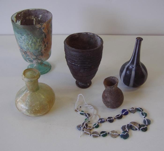 6 Roman Glass Objects, Vases, Breakers, Etc: Roman glass comprised of two footed beakers, a pear shaped vase, a squat-formed vessel, a miniature vessel and a glass necklace. Largest piece measuring 4.5" high. The smallest piece measuring 2" high