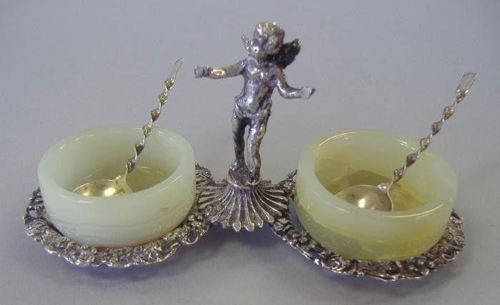German Silver Salt Cellar Stand & Sterling Spoons: German silver double salt stand with alabaster cellars. Figural winged cherub handle. The stand is marked 840. Comes with salt spoons, both marked sterling. Overall weight of the stand and spoons, 2.2