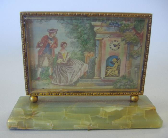 Flirt Fabsuisse Mechanical Desk Clock: Brass and glass picture frame desk clock showing a courting couple watching an angel hammering (when wound the angel hammers), within a tower. Mounted on an alabaster base. The back of the clock reads