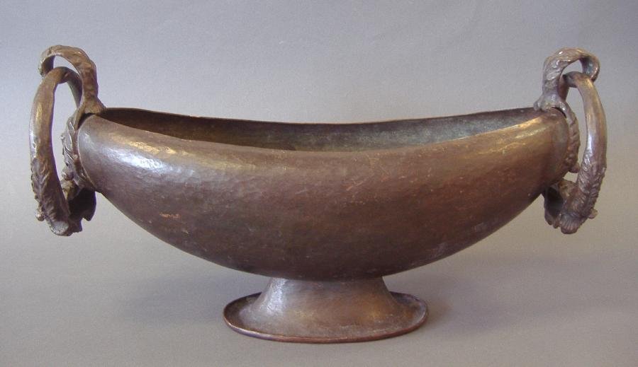Marie Zimmermann Hammered Copper Centerpiece Bowl: Marie Zimmermann ovoid footed centerpiece bowl of hammered copper. Leaf-formed ring handles with rams heads. Original dark brown patina. Measures 15.5" x 6" x 8" high to top of handle. NOTE: some pati