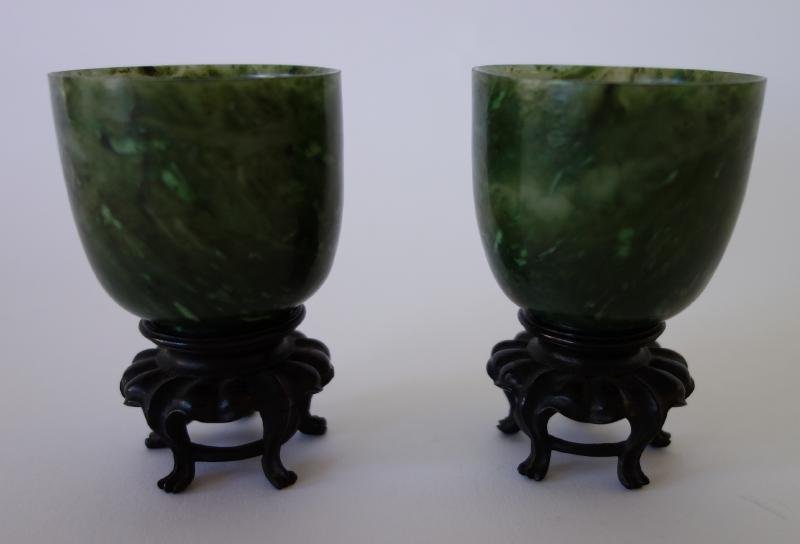 2 Chinese Spinach Jade Cups: A pair of Chinese spinach jade footed cups. Dark mottled stone with good translucency. Each measuring 2" high x 2" in diameter. With carved rosewood stands. NOTE: partial loss to rim of one wood stand