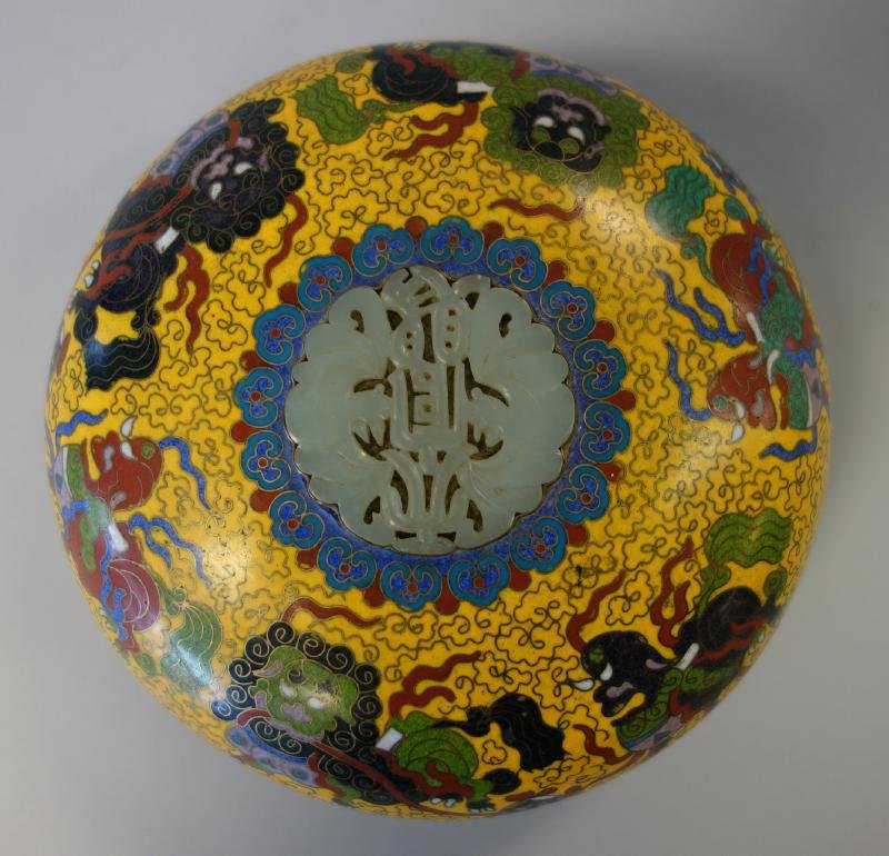 Chinese Cloisonne Enamel & Jade Box: A Chinese cloisonne enamel round presentation box. The lid inset with a carved jade medallion depicting two bats. The exterior decorated with a foo dog / lion motif. The inside as well as the exterior