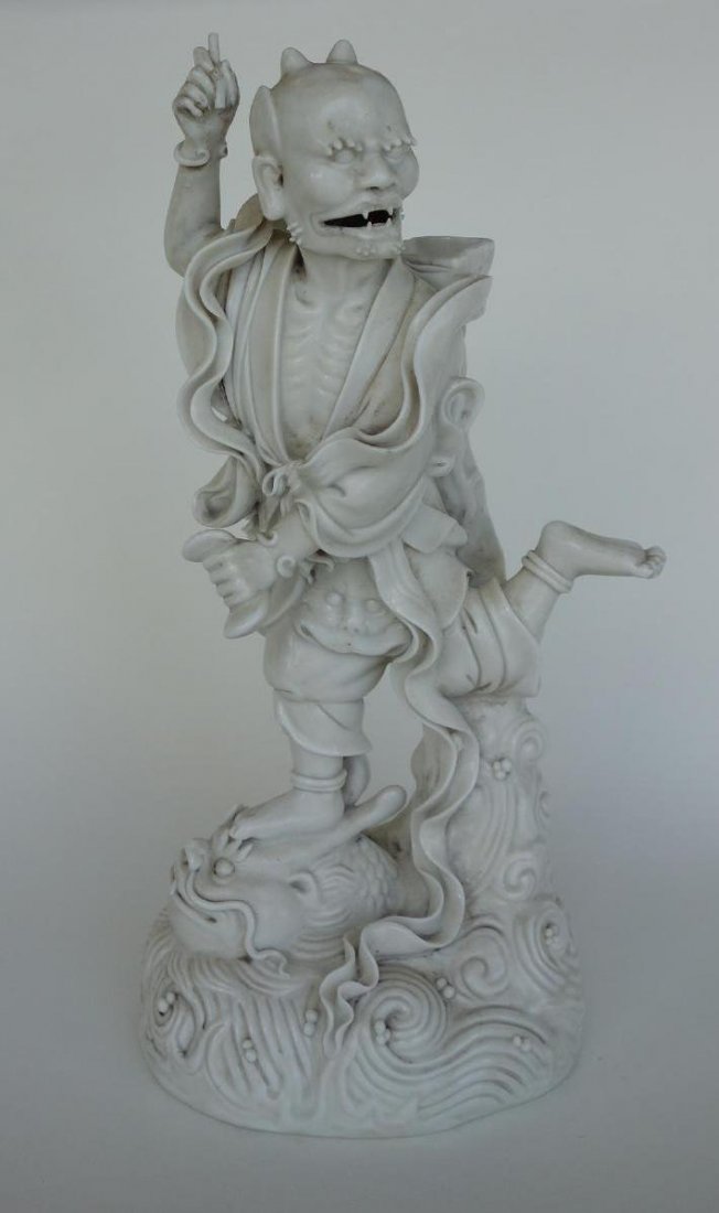 Chinese Blanc de Chine Horned Male Deity: A Chinese blanc de chine porcelain model of a horned male deity or devil. The clothed figure with horns is seen stomping on a mythological beast riding on top of waves. Chinese calligraphy on the bask