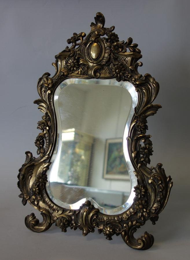 19thc Brass Table Top Vanity Mirror: A brass table-top vanity mirror. Late 19th / early 20th century. Embossed floral, swag and bird motif. Beveled mirror. Easel backed. Measures 17.25" high, 12" wide.
