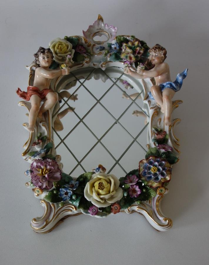 Sitzendorf Porcelain Figural Framed Mirror: A German porcelain framed mirror. Sitzendorf, Germany. Overall raised flower motif with part nude male figures flanking the mirror. Signed with the Sitzendorf cipher. NOTE: loss to one end of the figu