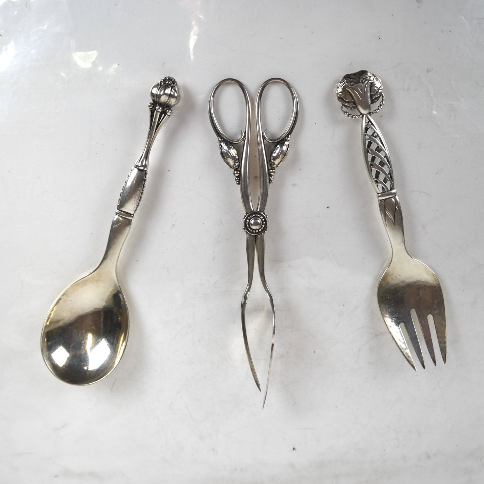 Georg Jensen Group: Tongs, Fork, Spoon: Tuli tongs #84, reticulated fork #18, reticulated spoon #38, .925 Sterling. [Largest: 8 inches]. 8.1 ozt. In good condition.