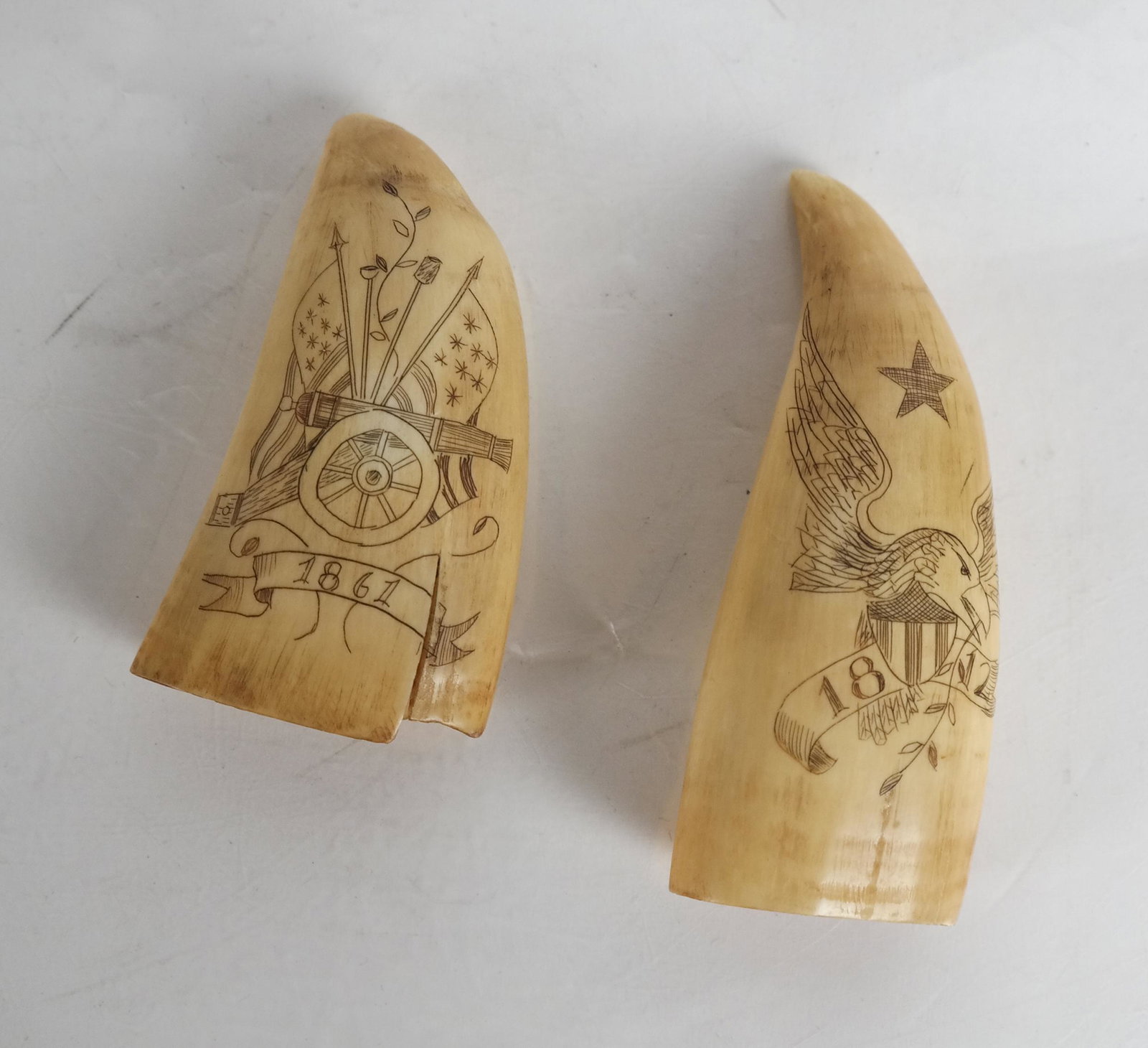 Two Scrimshaw Tooth Carvings, Dated 1812 & 1861: 1. Tooth dated 1861 depicting waving flags around a cannon [3 12/inches]. 2. The other, dated 1812, depicting an eagle atop a U.S. coat-of-arms with star atop [4 inches].